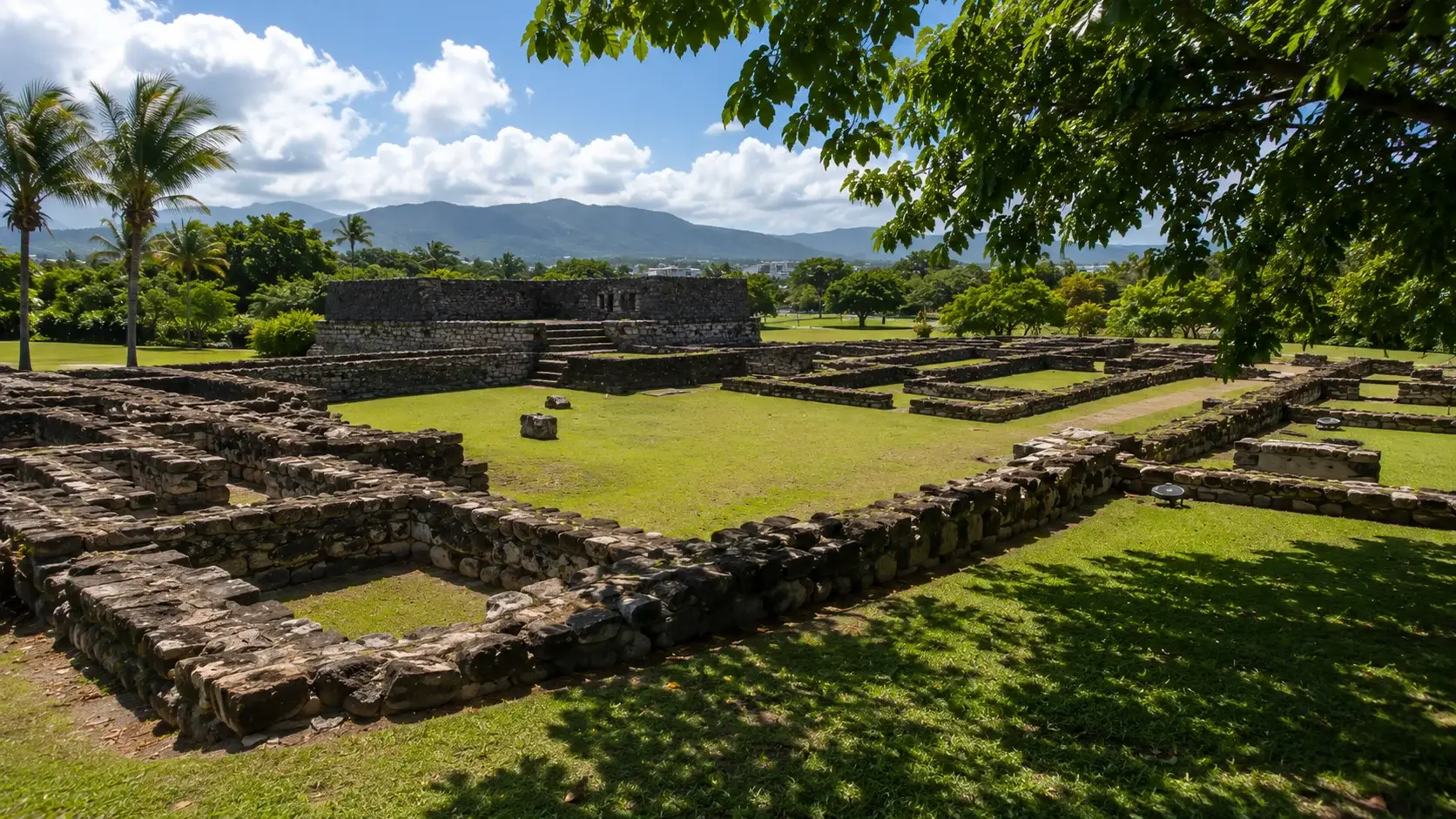 Ancient stone ruins at the Caparra Archaeological Site in Guaynabo, Puerto Rico, surrounded by tropical gardens with mountain views under a sunny blue sky.