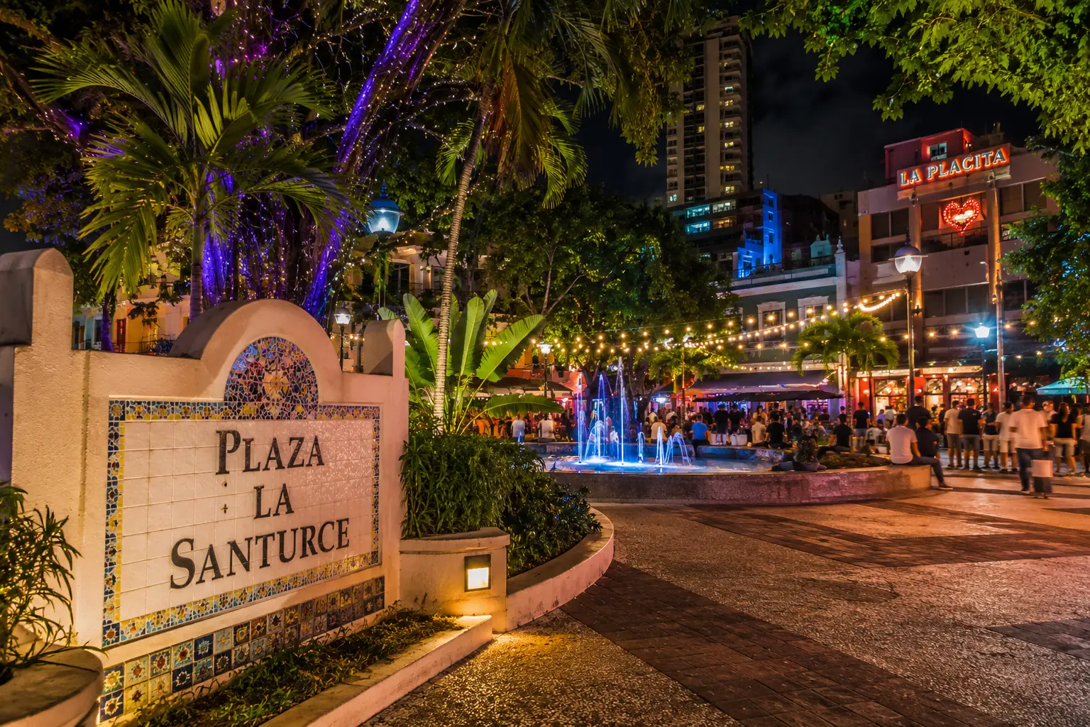 A stone plaza sign, palm trees and illuminated restaurants frame La Placita de Santurce at night in San Juan.