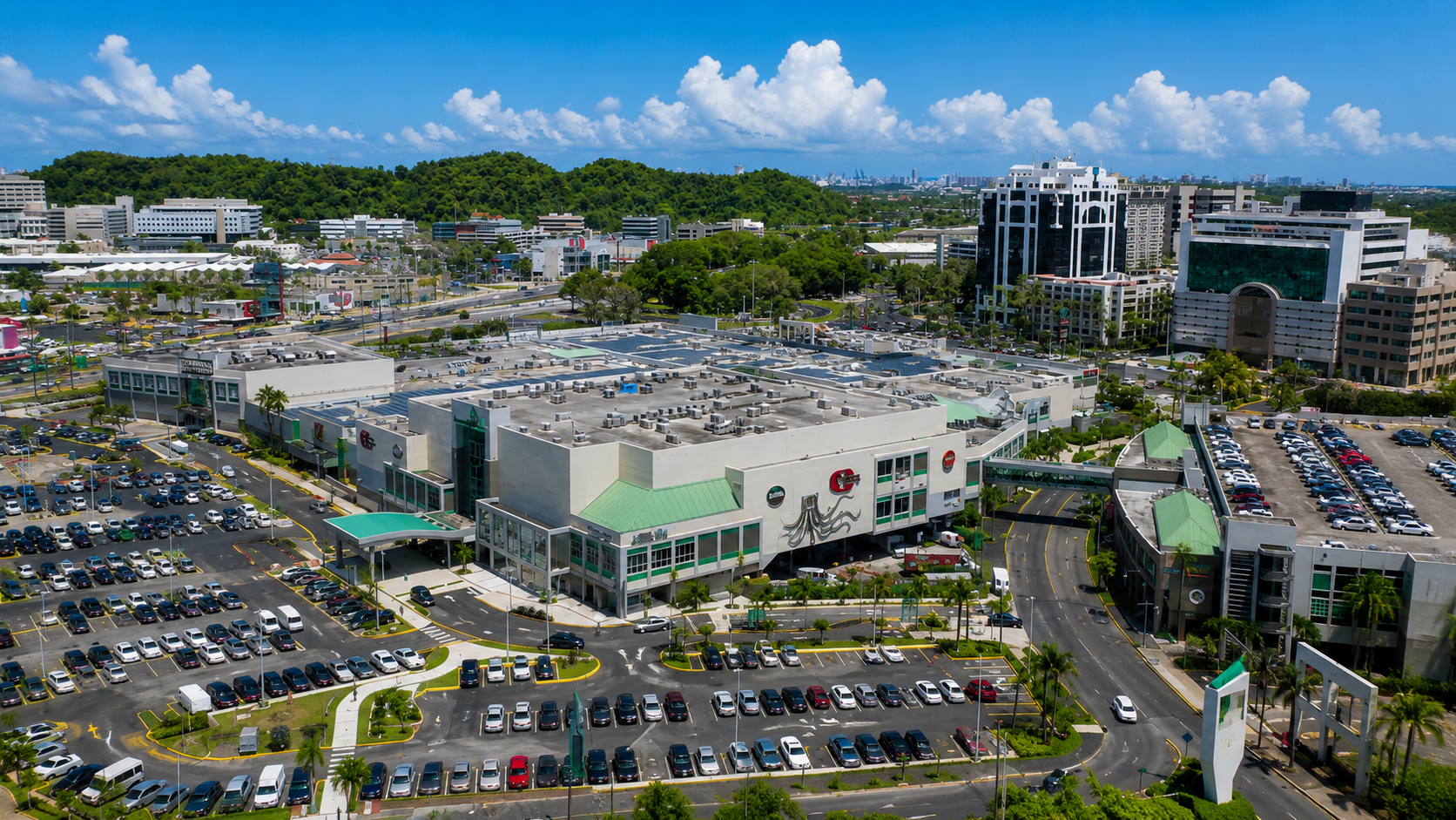 Aerial view of San Patricio Plaza in Guaynabo, Puerto Rico, featuring a modern shopping center, landscaped parking areas, office buildings, and the surrounding metropolitan skyline.