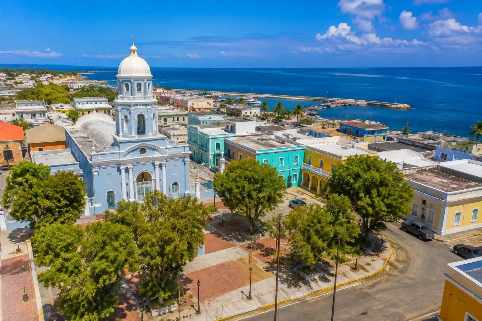 Aerial view of Aguadilla Pueblo in Puerto Rico, with a historic church, colorful buildings, shaded plaza, coastal streets, and blue ocean in the background.