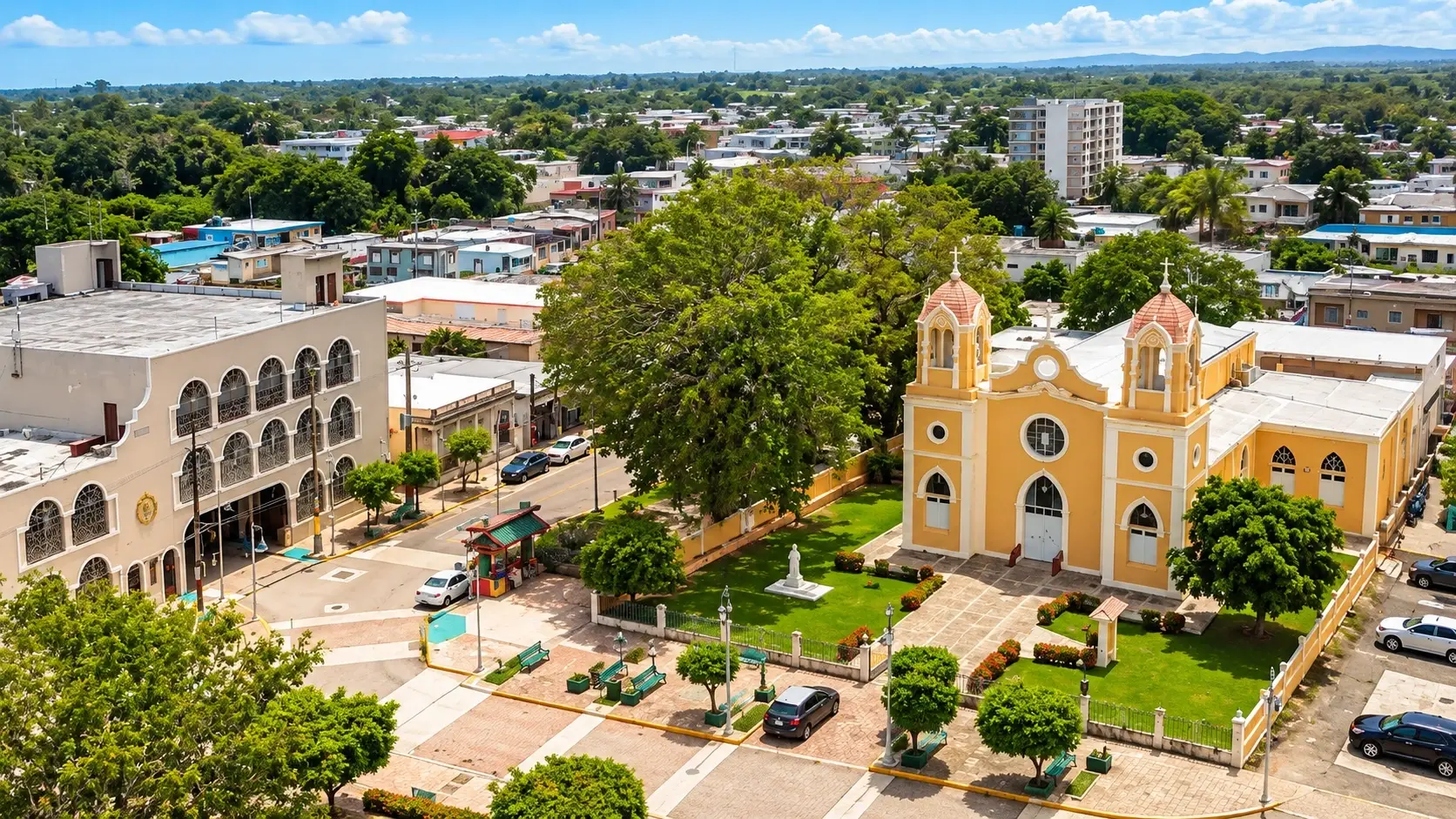 Aerial view of Añasco Pueblo in Puerto Rico, showing a historic church, landscaped plaza, colorful buildings, streets, and lush tropical surroundings.