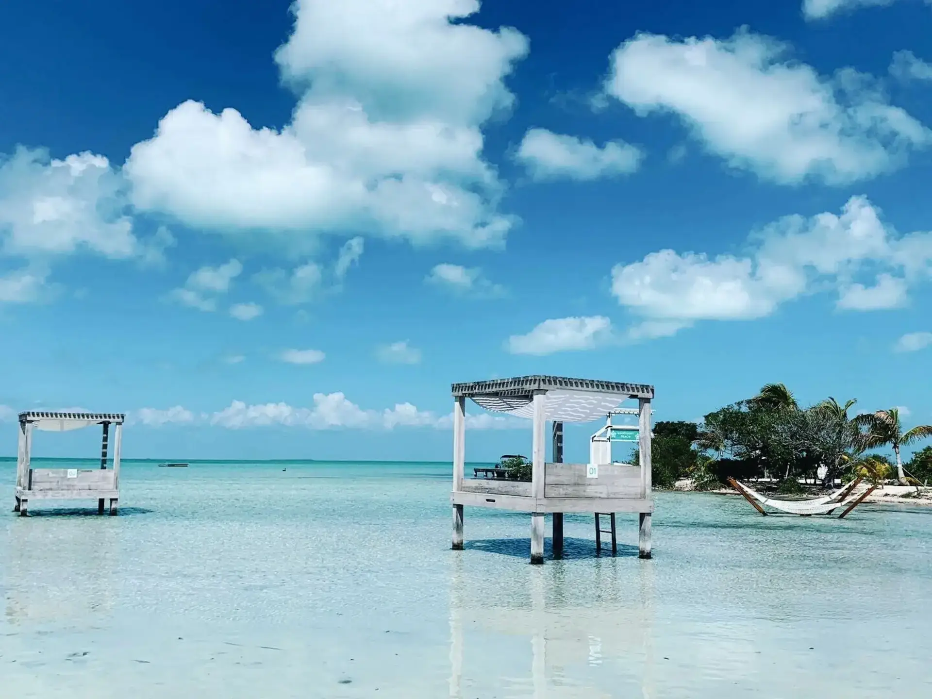 White wooden gazebos over turquoise shallows beside hammocks and a palm-lined beach in Belize