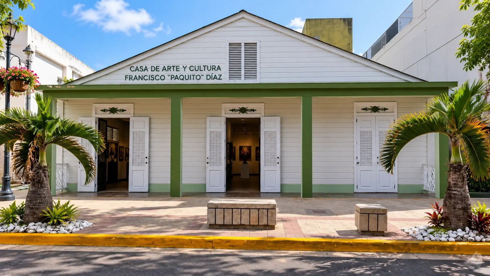 Exterior view representing Casa de Arte y Cultura Don Francisco “Paquito” Díaz in Aguas Buenas, Puerto Rico, with white wooden architecture, green trim, palm trees, and open gallery entrances.