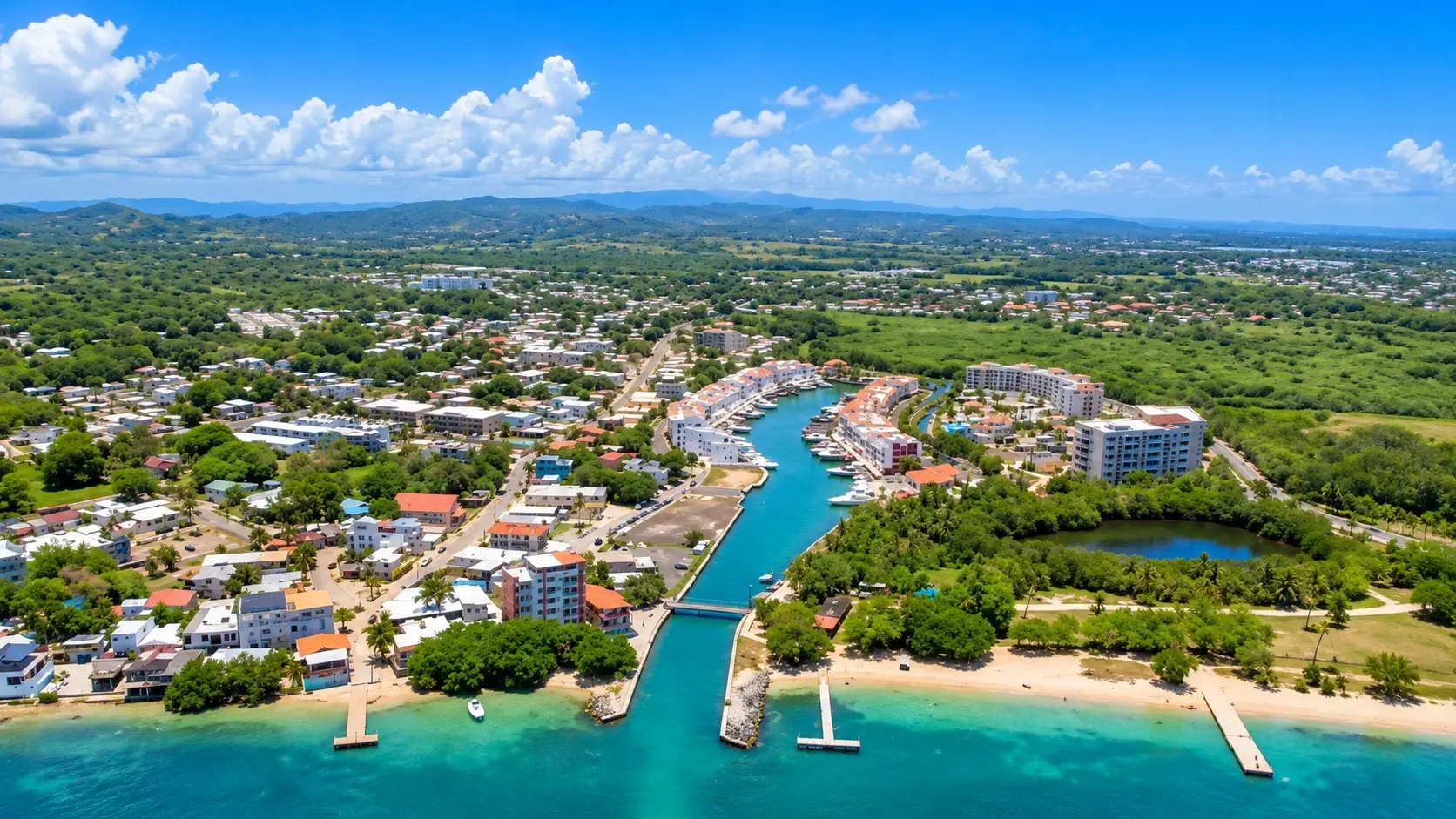 Aerial view representing Ceiba Baja in Aguadilla, Puerto Rico, with coastal residential development, waterfront buildings, roads, green land, tropical vegetation, and surrounding countryside.