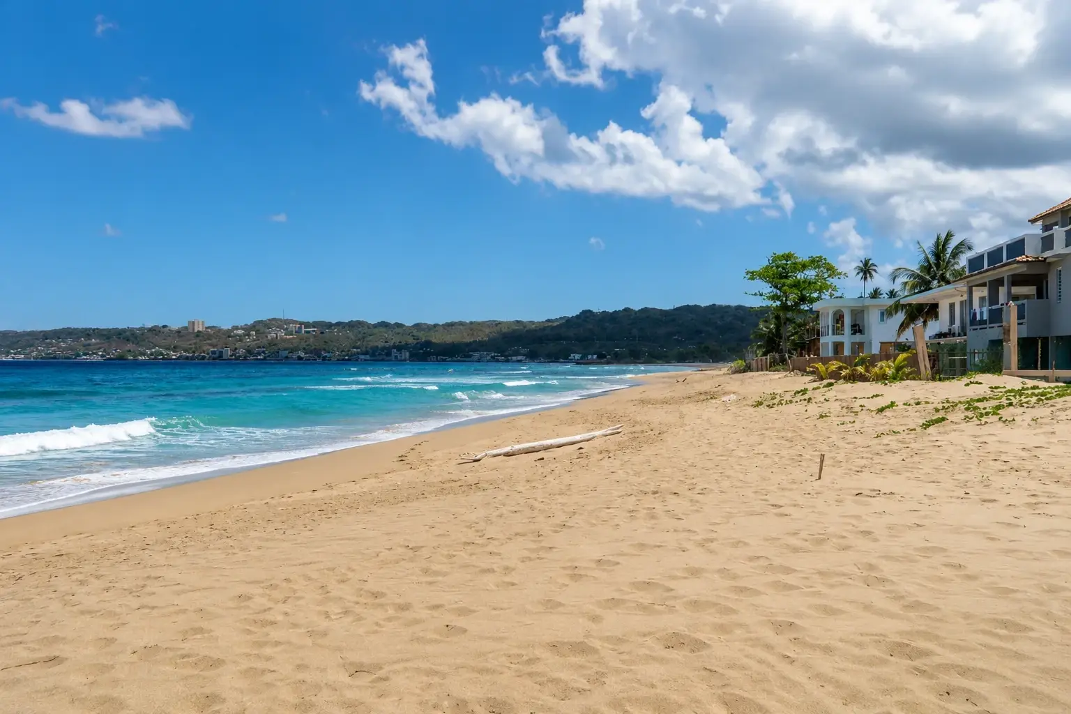 Wide sandy beach in Espinar, Aguada, Puerto Rico, with turquoise ocean waves, tropical palm trees, coastal homes, green hills, and a bright Caribbean sky.