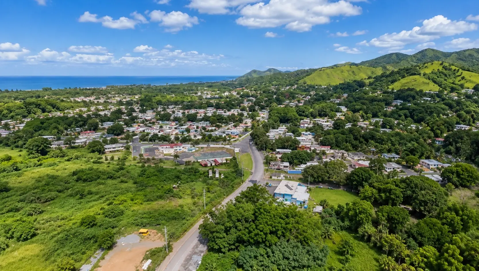 Aerial view representing Espino in Añasco, Puerto Rico, with residential communities, open land, local roads, tropical greenery, rolling hills, and distant ocean views.