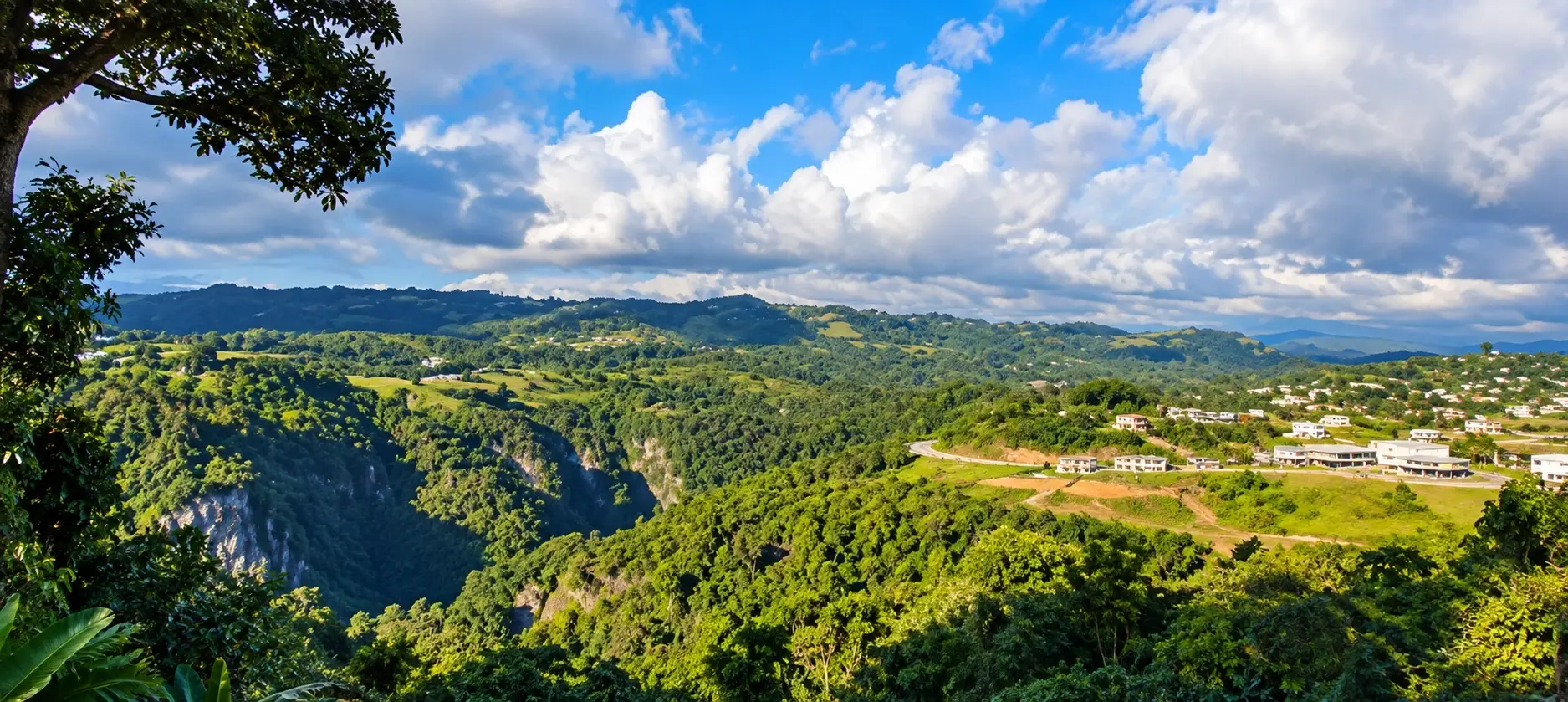 Panoramic view representing Llanos in Aibonito, Puerto Rico, with lush green mountains, residential areas, roads, development sites, and tropical countryside.