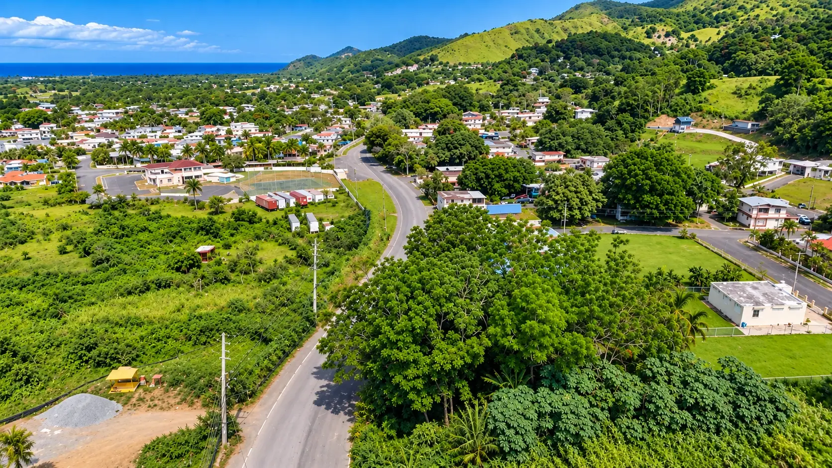 Aerial view representing Piñales Barrio in Añasco, Puerto Rico, with local roads, homes, lush tropical vegetation, rolling green hills, and distant coastal views.