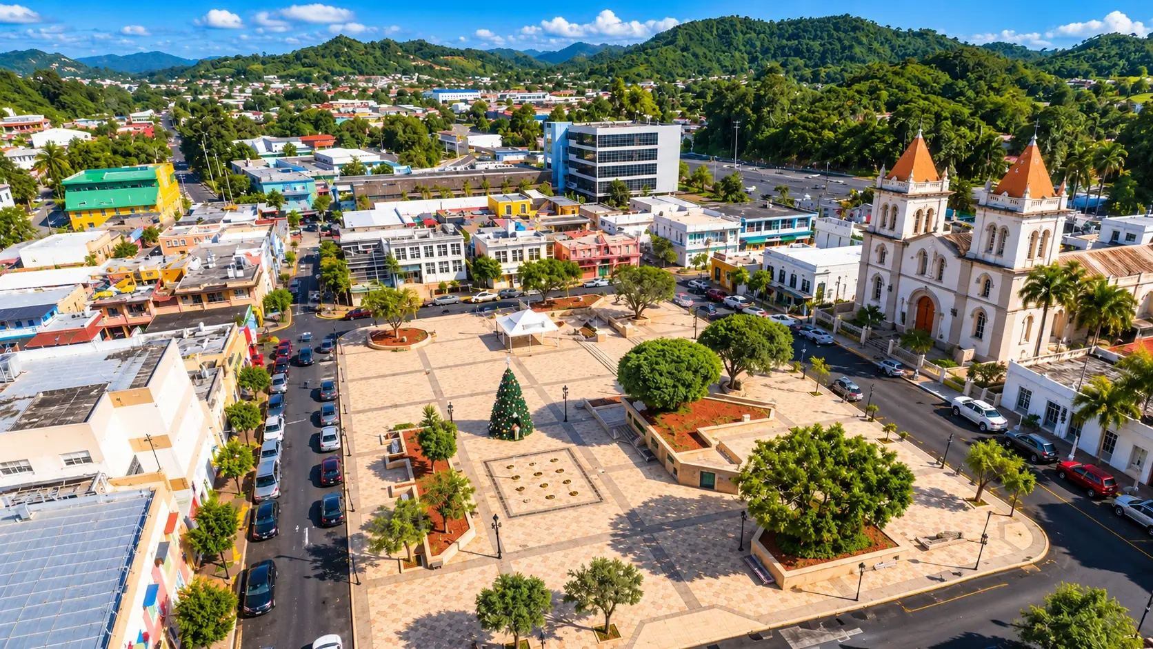 Aerial view representing Plaza de Recreo in Aibonito, Puerto Rico, with landscaped public spaces, surrounding buildings, streets, and green mountains.