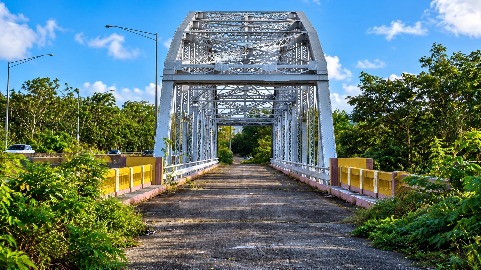 Historic Puente Salcedo in Añasco, Puerto Rico, featuring a steel truss structure, roadway, tropical vegetation, and bright blue sky.