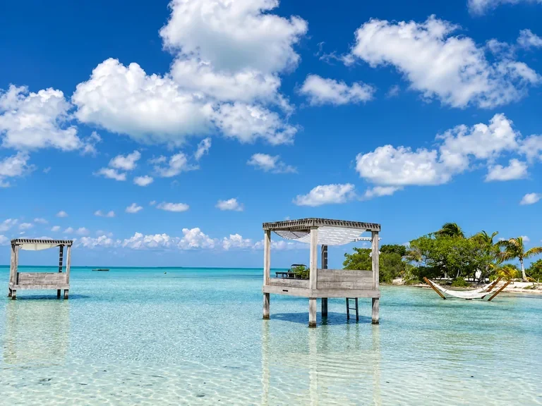 White wooden gazebos over turquoise shallows beside hammocks and a palm-lined beach in Belize