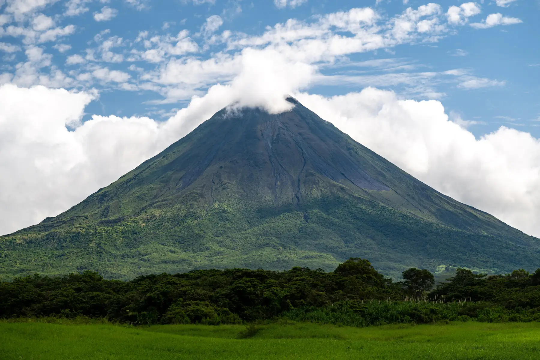 Arenal Volcano rising above green fields and forest beneath a partly cloudy blue sky in Costa Rica