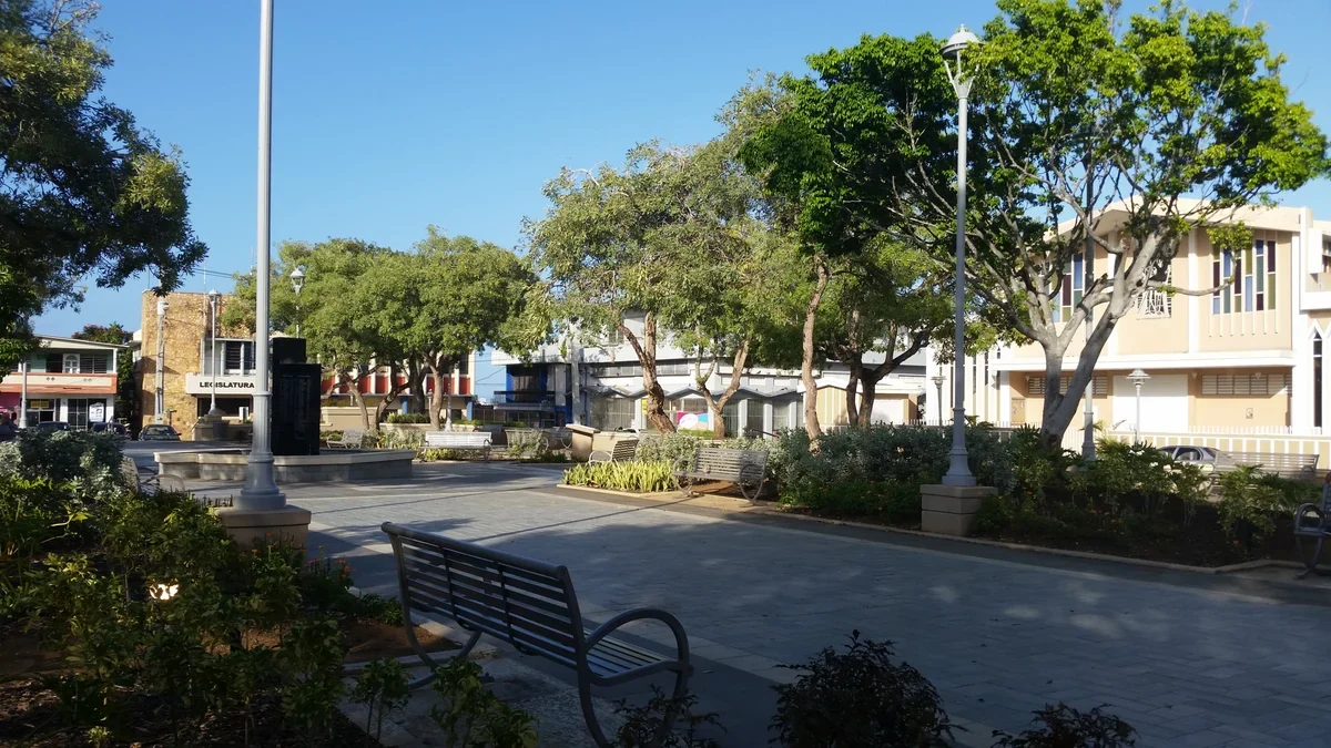 Benches, paved walkways, leafy trees and surrounding buildings at Luis Muñoz Marín town square in downtown Camuy, Puerto Rico.