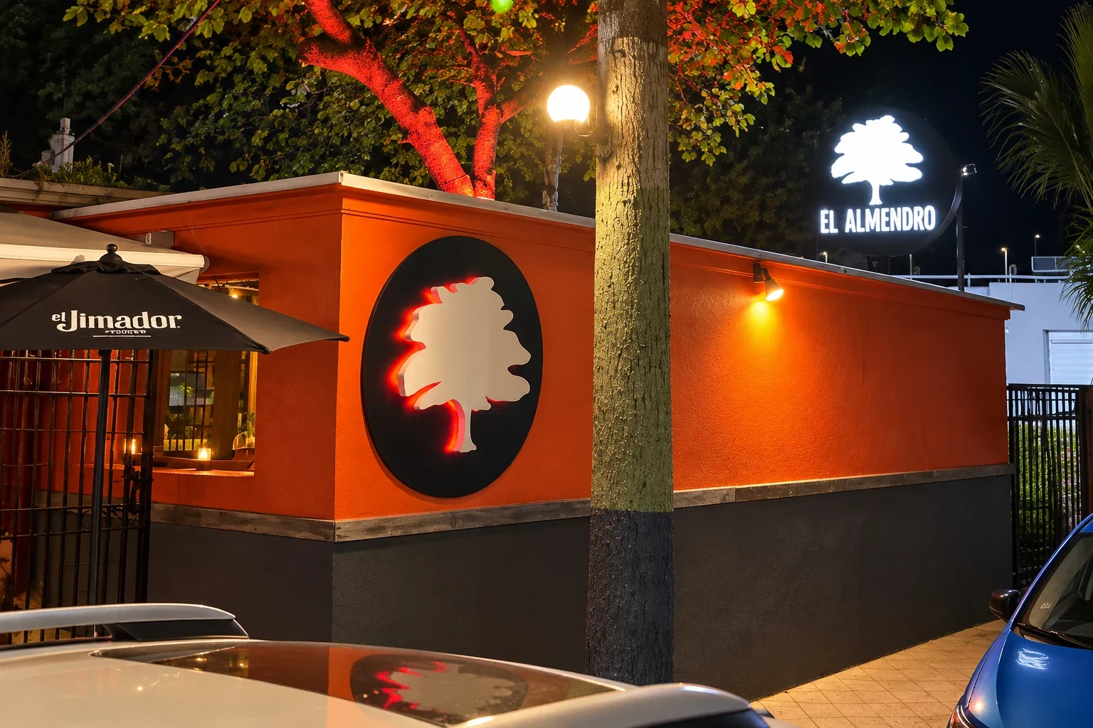 AI-enhanced image based on the El Almendro reference photo, showing its orange and dark-gray exterior, tree emblem, illuminated sign and nearby tree in Camuy, Puerto Rico.