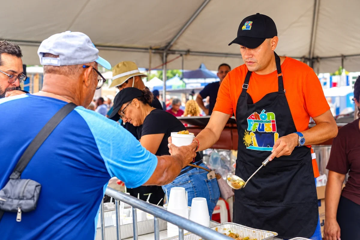 AI-enhanced photograph based on the supplied reference, showing a volunteer serving food to an attendee at Festival del Mondongazo in Camuy, Puerto Rico.