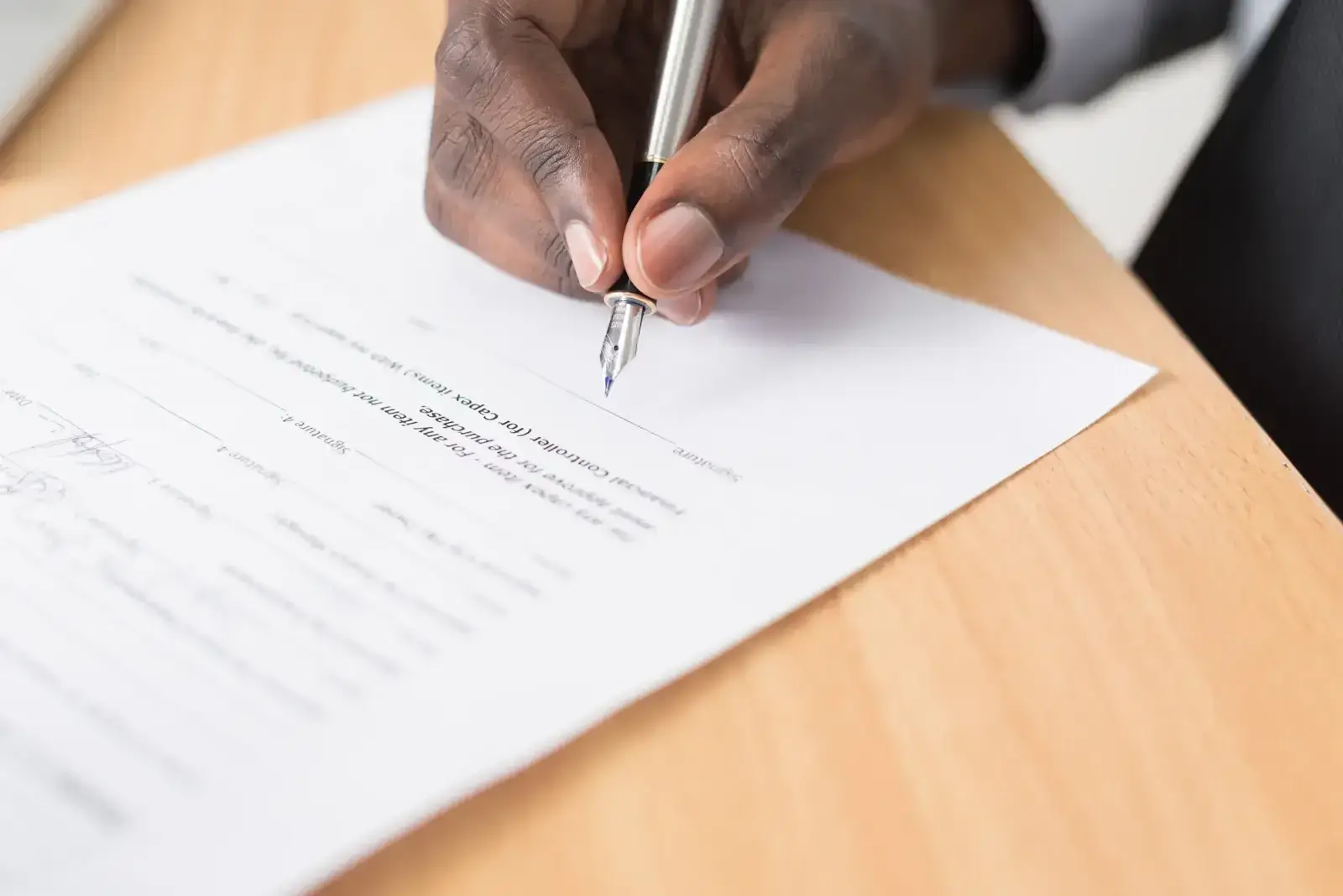Close-up of a person signing a printed document with a silver pen at a wooden desk.