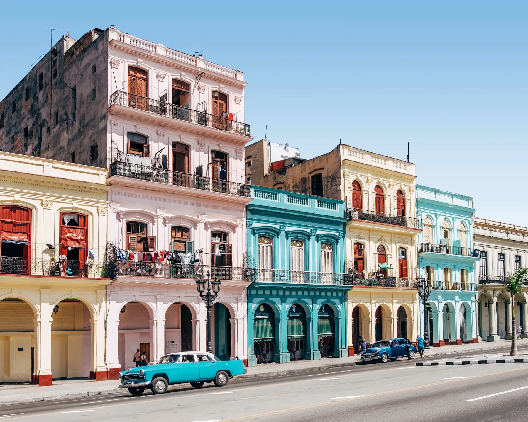 Colourful Havana buildings with balconies and arcades beside a road with classic cars under a blue sky