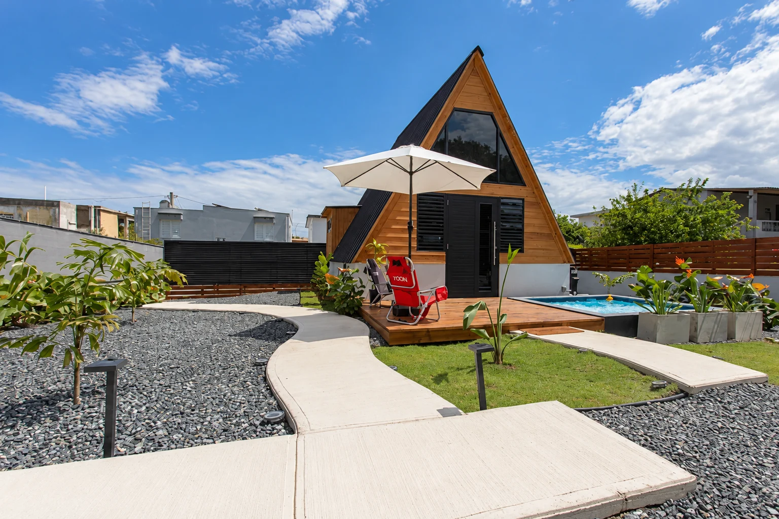 A-frame wooden cabin, winding concrete path, white patio umbrella, red lounge chair and small pool at Las Casitas in Camuy, Puerto Rico.