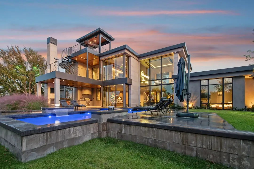 Modern Glass-Walled Home and Pool at Dusk A modern glass-walled house has illuminated rooms, a patio, and a swimming pool at dusk.
