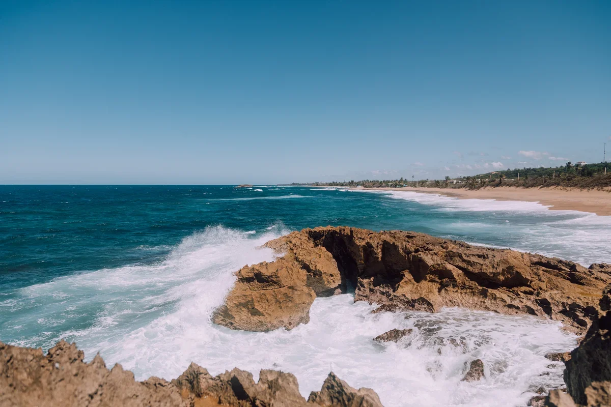 Atlantic waves break against golden coastal rocks at Peñón Amador in Camuy, Puerto Rico, with turquoise water, a sandy beach and clear blue sky.