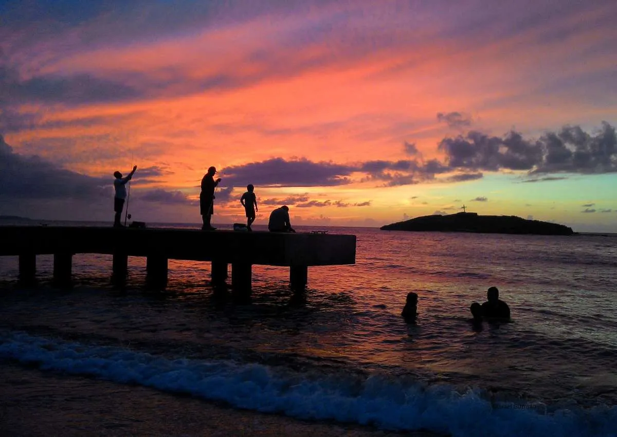 Pink, orange and purple sunset over the sea near Peñón Amador in Camuy, Puerto Rico, with people silhouetted on a pier and in the waves. Photo: Israel Guzmán.