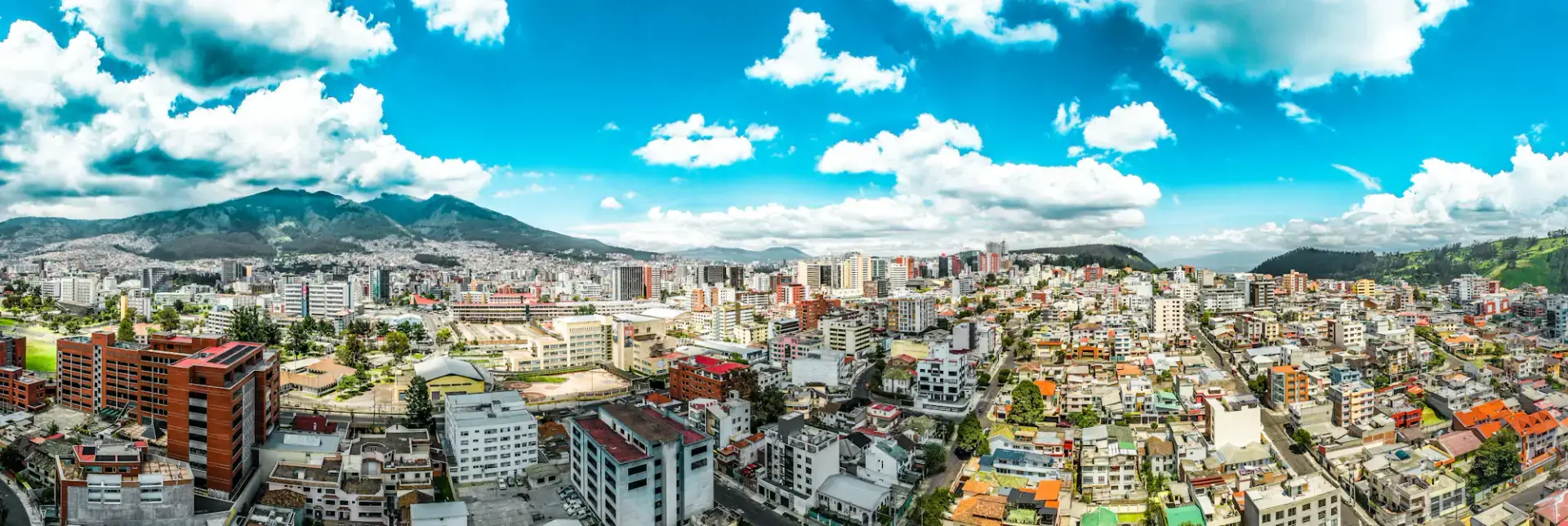 Quito city skyline and neighborhoods beneath green mountains and a bright cloud-filled sky