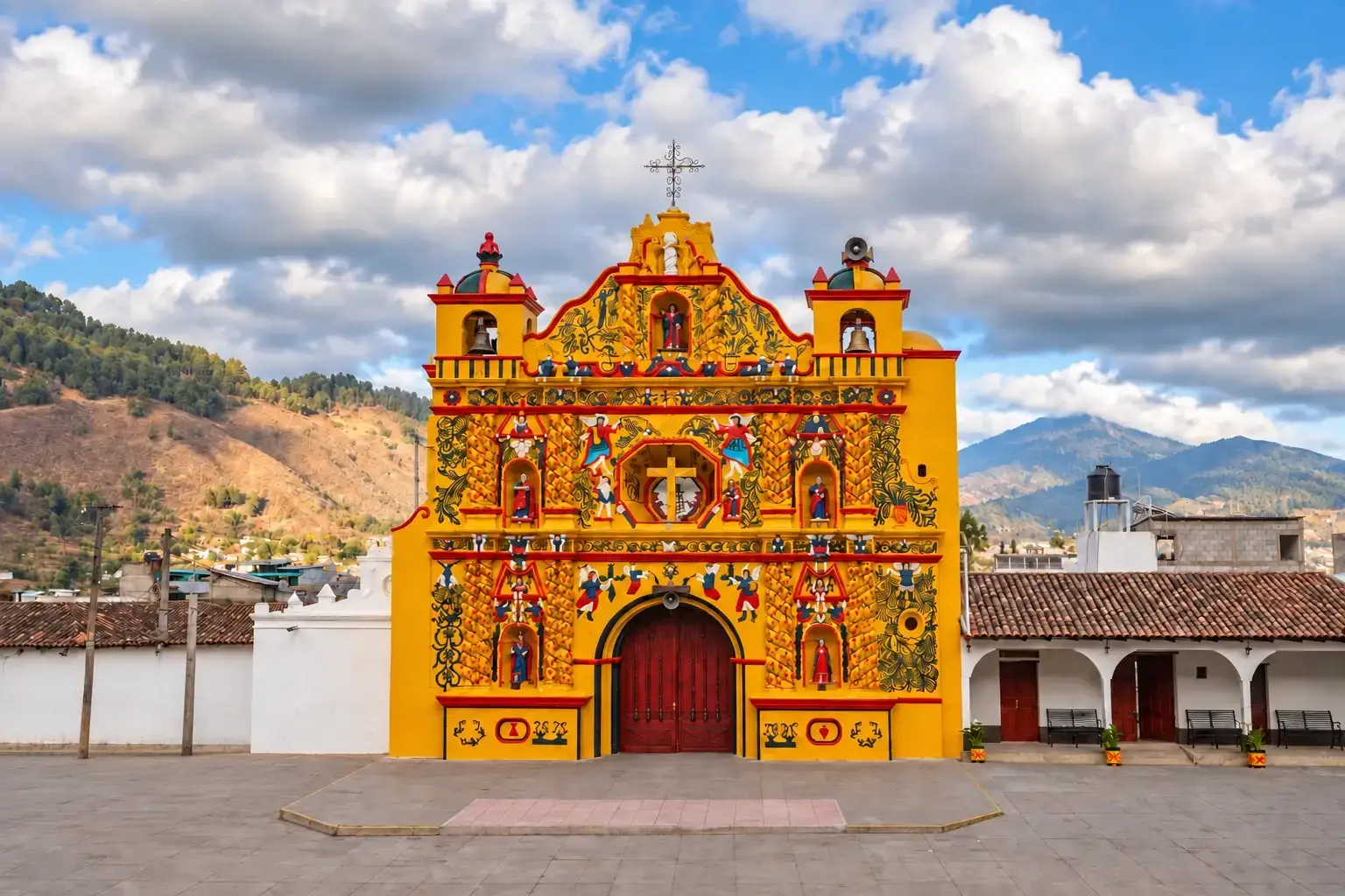 Centered yellow decorated San Andrés Xecul church with its cross against a cloudy blue sky, an empty plaza, tiled roofs and surrounding hills in Guatemala