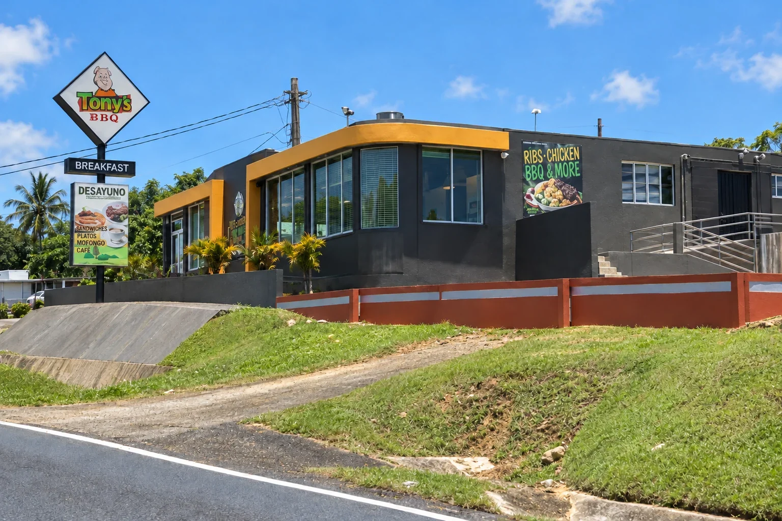 AI-enhanced image based on the user-supplied Tony’s BBQ reference photo, showing a dark gray restaurant façade with yellow trim, large windows, a roadside sign, access ramp and grassy verge in Camuy, Puerto Rico.